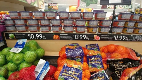 Canned goods stacked on shelves above bags of oranges and apples in a grocery store produce section. Prices visible.