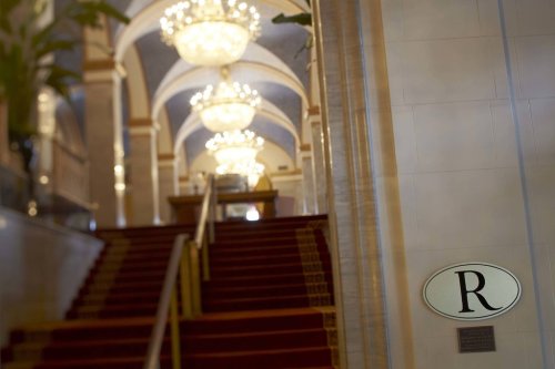 Elegant staircase with red carpet and chandeliers, featuring a sign with the letter "R" on the wall.