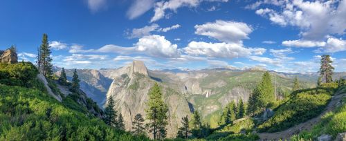 A panoramic view of a mountainous landscape with lush greenery, towering cliffs, and a bright blue sky with clouds.