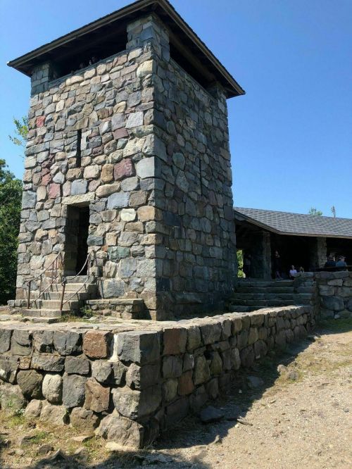 A tall stone tower with a flat roof, surrounded by a stone wall, under a clear blue sky. People are gathered nearby.