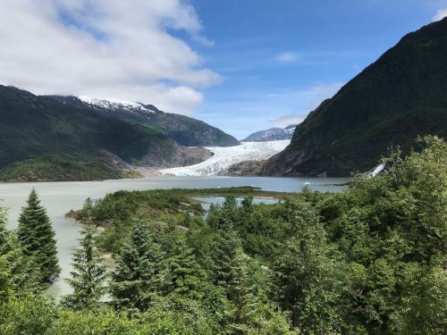 A stunning landscape featuring a glacier surrounded by mountains, lush greenery, and a serene lake under a blue sky.