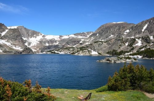 A serene mountain lake surrounded by rocky peaks and patches of snow, with green shrubs in the foreground.