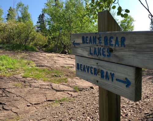 Wooden signpost with blue lettering pointing to Beane Bear Lakes and Beaver Bay, surrounded by greenery and rocky terrain.
