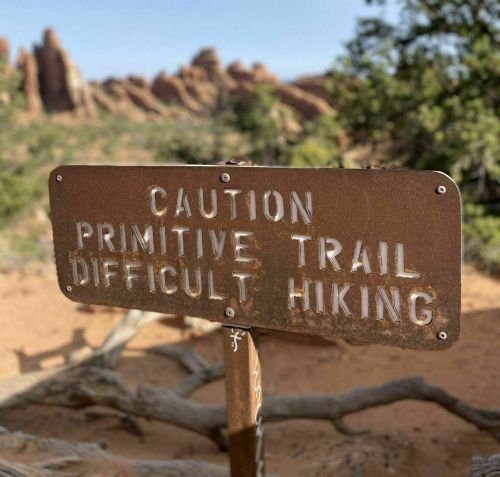 A weathered sign reading "Caution: Primitive Trail, Difficult Hiking" in a desert landscape with rocky formations.