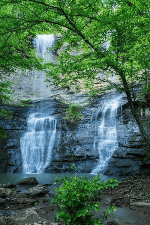 A serene waterfall cascades down rocky cliffs, surrounded by lush green trees and foliage.