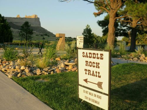 Sign for Saddle Rock Trail with scenic landscape and rock formations in the background, surrounded by greenery.