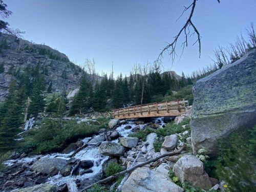 A wooden bridge spans a rocky stream surrounded by tall trees and rugged mountain terrain under a clear sky.