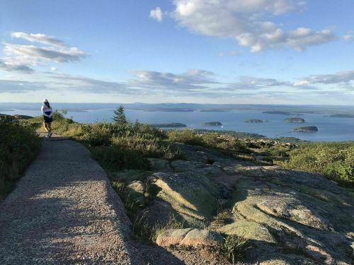 A person stands on a rocky path overlooking a scenic view of islands and water under a blue sky with clouds.