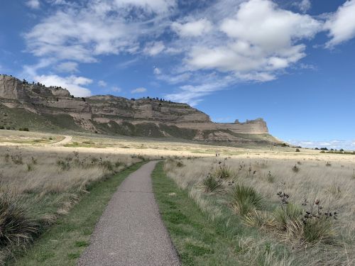 A winding path leads through tall grass towards rocky cliffs under a blue sky with scattered clouds.