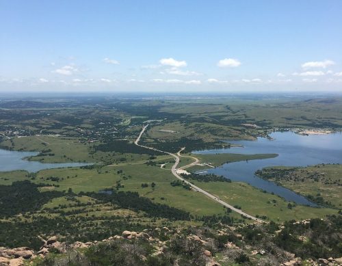 A panoramic view of rolling green hills, lakes, and a winding road under a clear blue sky.