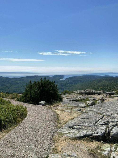 A scenic view from a mountain trail, featuring rocky terrain and lush green hills under a clear blue sky.