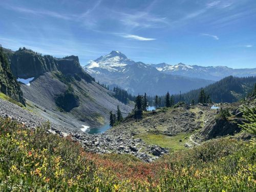 A scenic mountain landscape with lush greenery, rocky terrain, and snow-capped peaks under a clear blue sky.