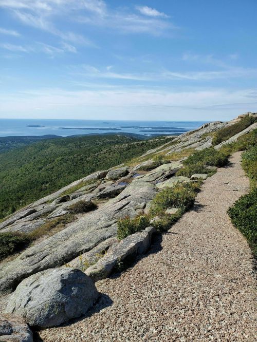 A rocky trail winds along a mountain, overlooking a scenic view of green hills and a blue ocean in the distance.