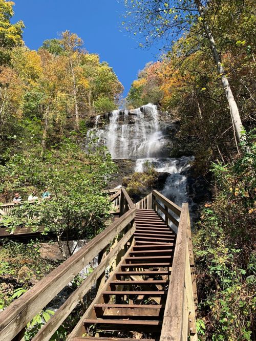 A wooden staircase leads to a waterfall surrounded by colorful autumn foliage under a clear blue sky.