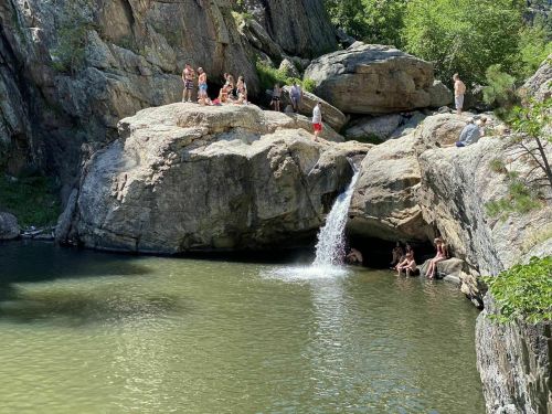 A scenic rocky area with a small waterfall flowing into a green pool, surrounded by people enjoying the outdoors.