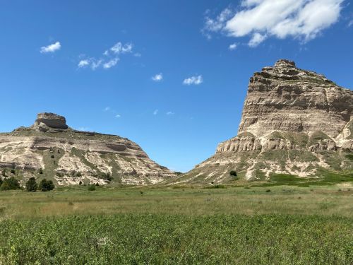 Two large rock formations rise above a green field under a blue sky with scattered clouds.