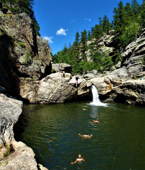 A scenic swimming spot with people enjoying the water, surrounded by rocky cliffs and lush greenery under a clear blue sky.