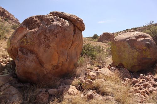 Two large boulders sit among dry grass and rocky terrain under a clear blue sky.