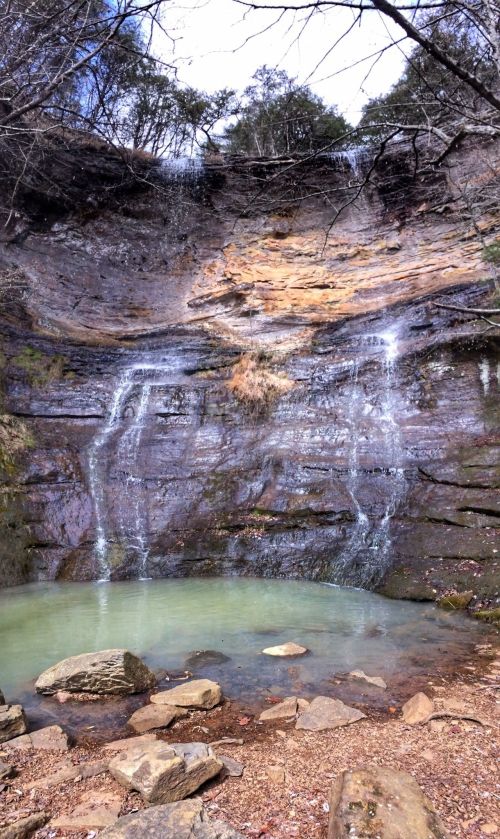 A serene waterfall cascading over rocky cliffs into a calm green pool, surrounded by trees and natural scenery.