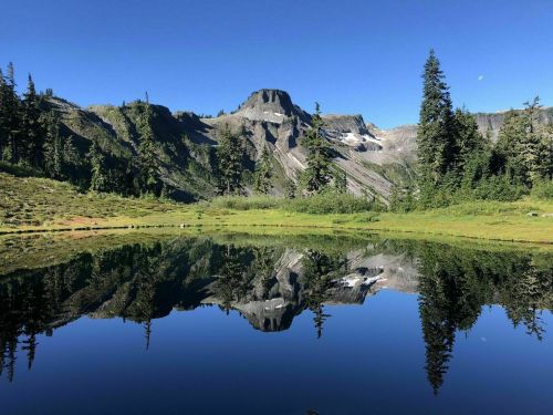 A serene mountain landscape reflected in a calm lake, surrounded by lush greenery and clear blue skies.