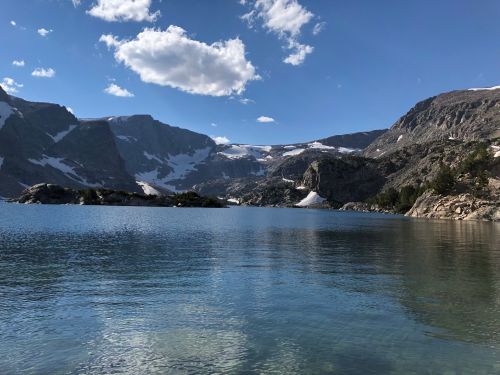 A serene mountain lake surrounded by rocky cliffs and snow-capped peaks under a blue sky with fluffy clouds.