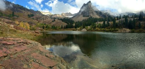 A serene mountain lake surrounded by rocky shores, lush trees, and towering peaks under a partly cloudy sky.