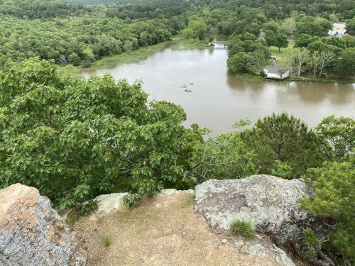 Aerial view of a lush green landscape with a calm lake and kayakers, surrounded by trees and rocky terrain.