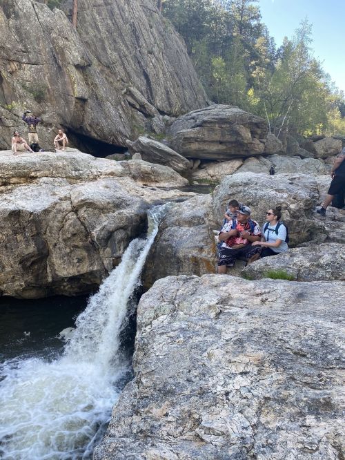 A group of people relax by a rocky riverbank with a waterfall, surrounded by trees and cliffs.