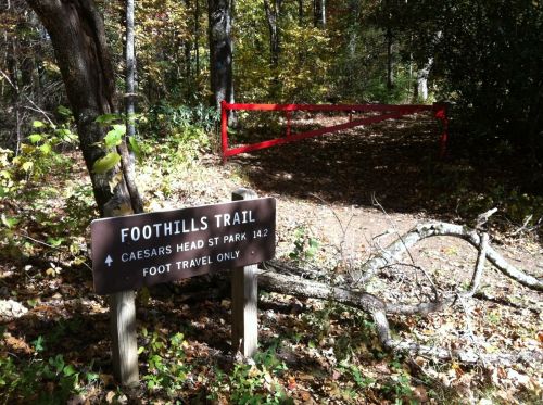 Sign for Foothills Trail at Caesars Head State Park, with a red gate in the background and a forested path ahead.