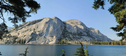 A serene lake surrounded by tall trees, with a large rocky mountain rising in the background under a clear blue sky.