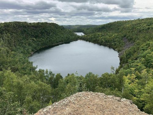 A serene lake surrounded by lush green forests under a cloudy sky, viewed from a rocky overlook.