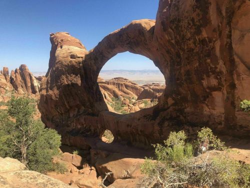 A natural rock formation with a large arch, surrounded by red rock formations and sparse vegetation under a clear blue sky.