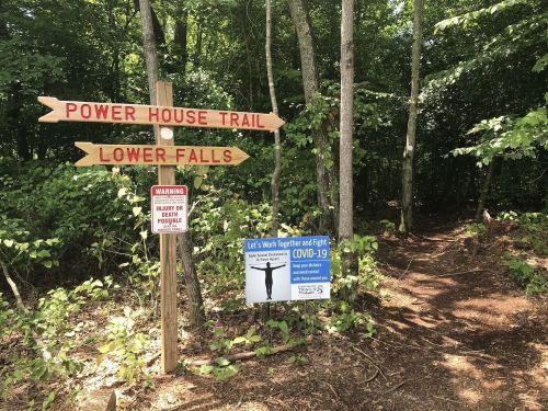 Signpost indicating trails for Power House and Lower Falls, with a COVID-19 safety reminder nearby in a wooded area.