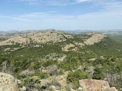 A panoramic view of rolling hills and rocky terrain under a clear blue sky, with lush greenery in the foreground.