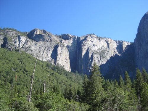 A towering granite cliff with a waterfall, surrounded by lush green trees under a clear blue sky.