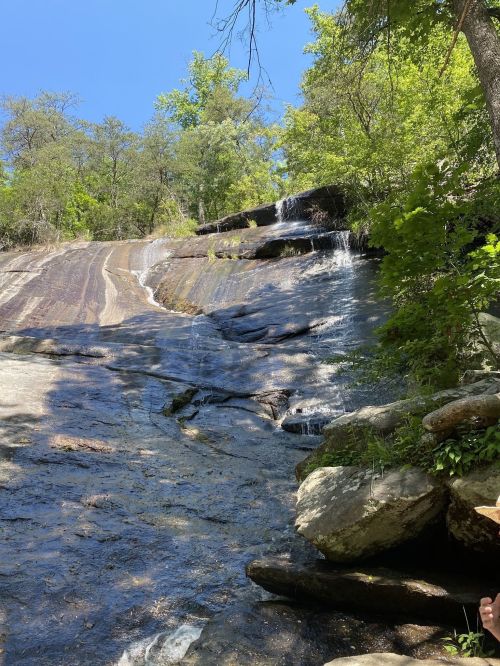 A serene waterfall cascading down a rocky slope, surrounded by lush green trees under a clear blue sky.