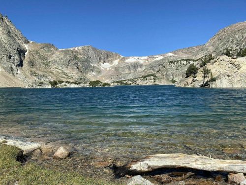 A serene lake surrounded by mountains under a clear blue sky, with rocky shores and a log in the foreground.