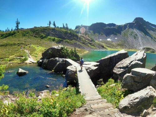 A person walks on a stone bridge over a clear lake, surrounded by lush greenery and mountains under a bright blue sky.