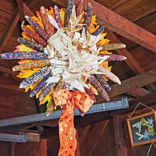 A decorative wreath made of colorful corn, dried leaves, and an orange ribbon, hanging in a rustic wooden setting.