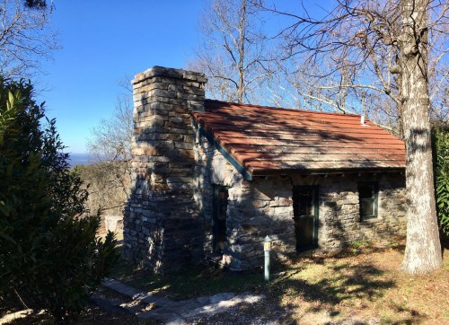 A small stone cottage with a red roof, surrounded by trees and a clear blue sky in the background.