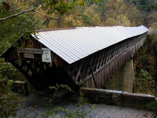 A rustic covered bridge with a sloped roof, surrounded by trees and autumn foliage, featuring a weight limit sign.