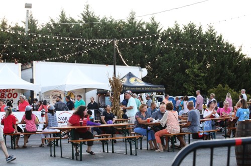 A lively outdoor event with people dining at picnic tables, food trucks, and festive string lights in the background.