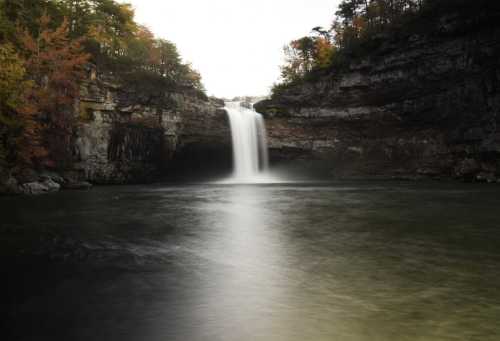 A serene waterfall cascades into a calm pool, surrounded by rocky cliffs and autumn foliage.