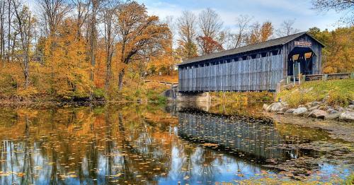 A wooden covered bridge reflects in a calm river surrounded by autumn foliage and colorful leaves.