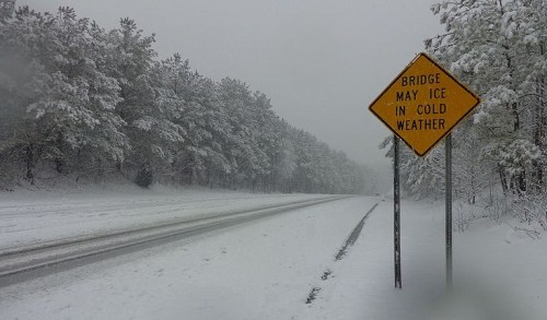 A snowy road with a warning sign that reads "Bridge May Ice in Cold Weather," surrounded by trees.