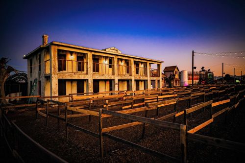 Abandoned two-story building at dusk, surrounded by wooden fences and a clear sky transitioning to night.