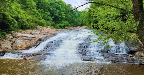 A serene waterfall cascades over rocky ledges, surrounded by lush green trees and a calm river below.