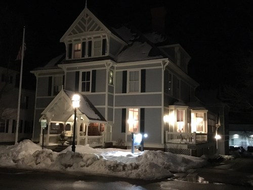 A large, historic house at night, with snow in front and warm lights glowing from the windows.