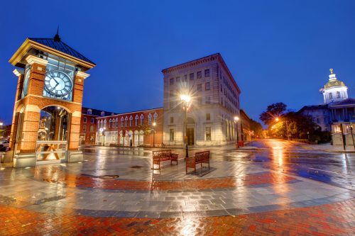 A quiet town square at dusk, featuring a clock tower, benches, and historic buildings reflecting wet pavement.