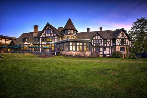 A large, elegant Tudor-style mansion with a grassy lawn and a sunset sky in the background.